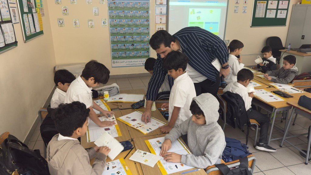 معلم يوجّه طلابًا أثناء نشاط تعاوني جماعي في الصف مع دفاتر عمل تفاعلية