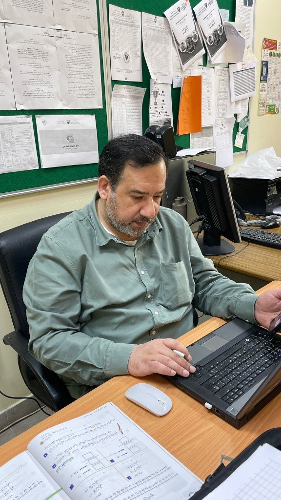 أ. فاضل يحيى أثناء العمل على الحاسوب والمتابعة الإدارية أمام لوحة إعلانات صفية