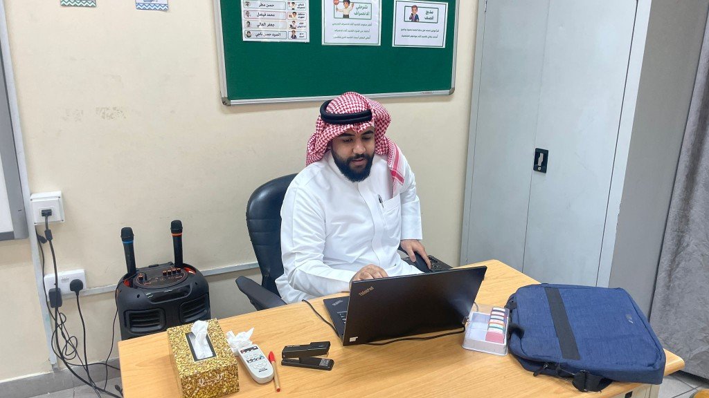 أ. محمد النجار أثناء العمل على الحاسوب والمتابعة الإدارية أمام لوحة إعلانات صفية