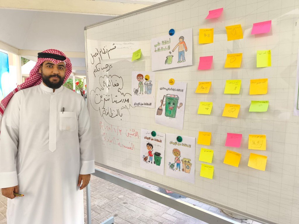 أ. محمد النجار بجانب لوحة فعالية مدرستنا تتزين بالنظافة في الفناء المدرسي